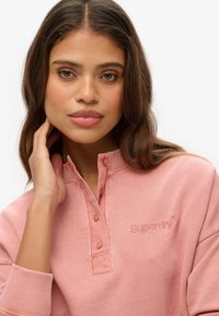 Sudadera de color rosa con cuello henley, que cuenta con tres botones y una tela texturizada. Logo bordado de "Superdry" en el pecho.