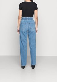 Žena stojící zády, oblečená v vysokých světle modrých kalhotách paperbag, černém přiléhavém tričku s krátkým rukávem a černých botách na podpatku.