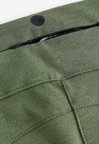 Zeleni materijal s džepom na zip, s okruglim crnim logom. Vidljiv je teksturirani tkanje i šivani detalji.