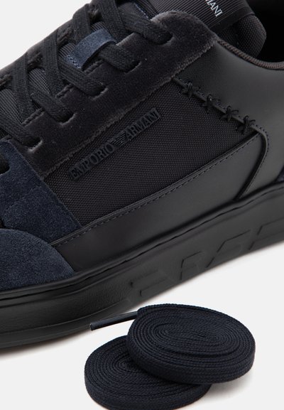 Μαύρο sneaker Emporio Armani με πάνελ από σουέτ και δίχτυ, λεπτομερείς ραφές και ένα επιπλέον ζευγάρι μαύρα σπιράλ κορδόνια σε λευκό φόντο.