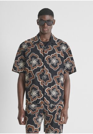 Homme portant des lunettes de soleil noires, une chemise à manches courtes noire à boutons et un short avec de grands motifs floraux marron et blanc.