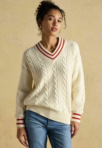 Kreemikaalune kootud kampsun V-kaelusega, millel on nöörmustrit ja punaste triipudega aktsendid kaeluses ja varrukatel. Koos siniste teksadega.