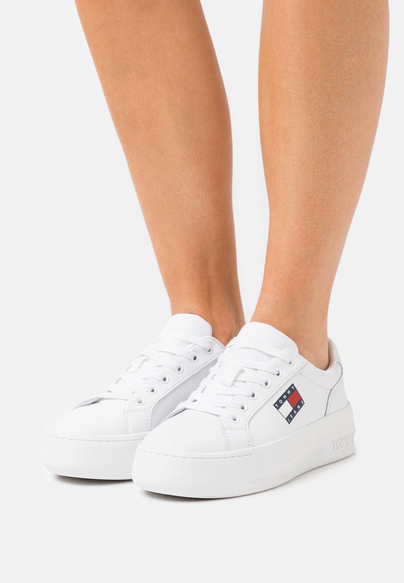 Pieds portant des baskets basses blanches avec des lacets blancs et un logo Tommy Jeans sur le côté, debout sur un fond clair uni.