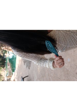 Türkise Haarbürste mit spiralförmigem Design und langen Zähnen, in einer Hand gehalten, neben langen, dunklen Haaren; strukturierter weißer Pullover sichtbar.