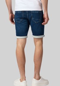 Dżinsowe szorty w ciemnoniebieskim kolorze z podwiniętymi mankietami, z dwiema tylnymi kieszeniami i dopasowanym krojem. Noszone z białymi sneakersami i prostą bluzką.