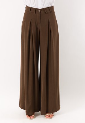 Pantaloni marroni a vita alta e gamba larga con pieghe frontali e chiusura con bottoni, indossati con una camicia bianca e sandali beige con punta aperta.
