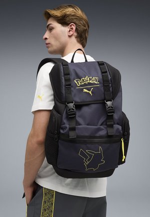 Młody mężczyzna noszący czarno-szary plecak w tematyce Pokémon z żółtym konturem Pikachu i logo Puma, ubrany w białą koszulę i szare spodnie.