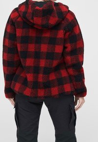 Czerwona i czarna kurtka polarowa w kratę z kapturem, wykonana z miękkiego, teksturowanego materiału o luźnym kroju. Widoczne szwy i zaokrąglony dolny brzeg.