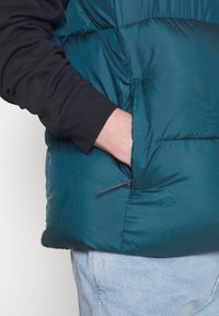 Türkisfarbene Steppjacke mit strukturiertem Material, ausgestattet mit einer Reißverschlusstasche. Schwarzer Ärmel sichtbar mit einem gerippten Bündchen; getragen über hellen Denim-Hosen.