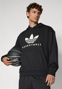 Fekete kapucnis pulóver nagy fehér "BASKETBALL" logóval és három csíkkal, fekete kosárlabdával párosítva.
