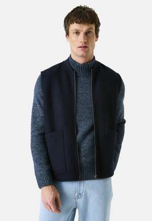 Marineblauw mouwloos vest met rits aan de voorkant, gemaakt van een wolmix, met twee voorzakken, gelaagd over een gestructureerde blauwe turtleneck trui. Lichtblauwe jeans.