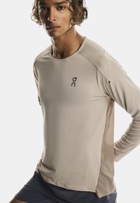 Maglietta sportiva a maniche lunghe di un beige chiaro, con un frontale testurizzato e pannelli laterali in rete. Include un piccolo logo sul petto.