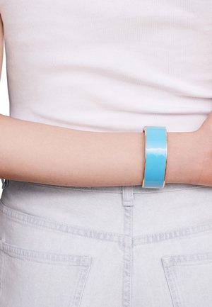 A wide, glossy turquoise bangle with a smooth finish and gold-tone edges, worn on a light-skinned arm against a white tank top.