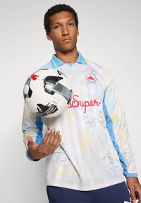 Camicia a maniche lunghe in tessuto leggero con colletto blu; presenta motivi floreali e un logo rosso. Palla da calcio in bianco e nero tenuta in mano.