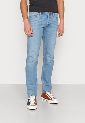 Mand iført lyseblå slim-fit jeans, en sort skjorte og tofarvede sneakers med blå og brune paneler samt hvide snørebånd.