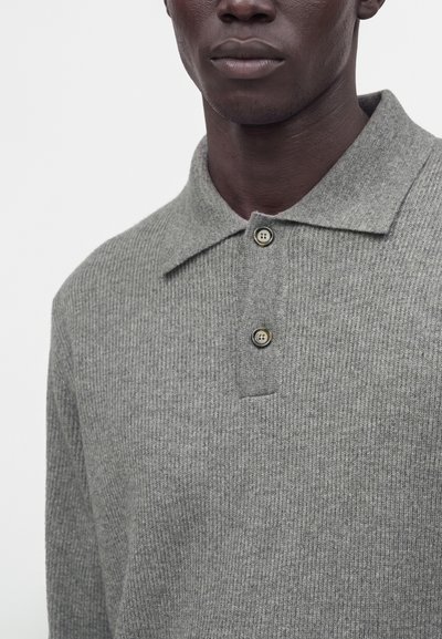 Homme portant un polo en maille côtelée grise avec deux boutons beiges, montré du bas du visage au milieu du torse sur un fond uni.