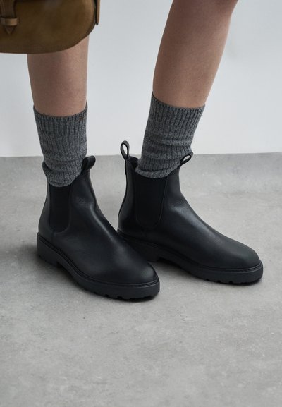 Zwarte leren Chelsea-boots met een ronde neus, elastische zijpanelen en aantrekbanden. Gecombineerd met grijze geribde sokken. Platte rubberen zool zichtbaar.