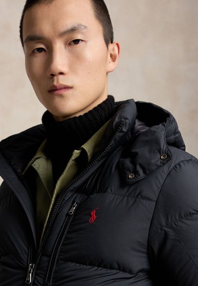 Young man wearing a black puffer jacket with a red logo, black turtleneck sweater, and olive green shirt against a plain beige background.