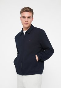 Sötétkék, cipzáros dzseki hegyes gallérral, oldalsó zsebekkel, rugalmas mandzsettákkal és kis logóval a mellrészen. Simább pamut anyag.