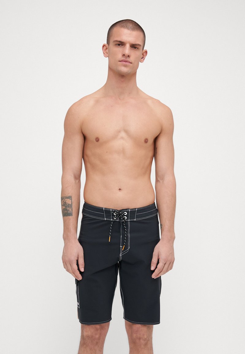 Shorts de bain noirs avec des coutures blanches, taille élastique avec cordon de serrage ajustable, poche latérale et coupe au genou ; fabriqués en matériau léger.