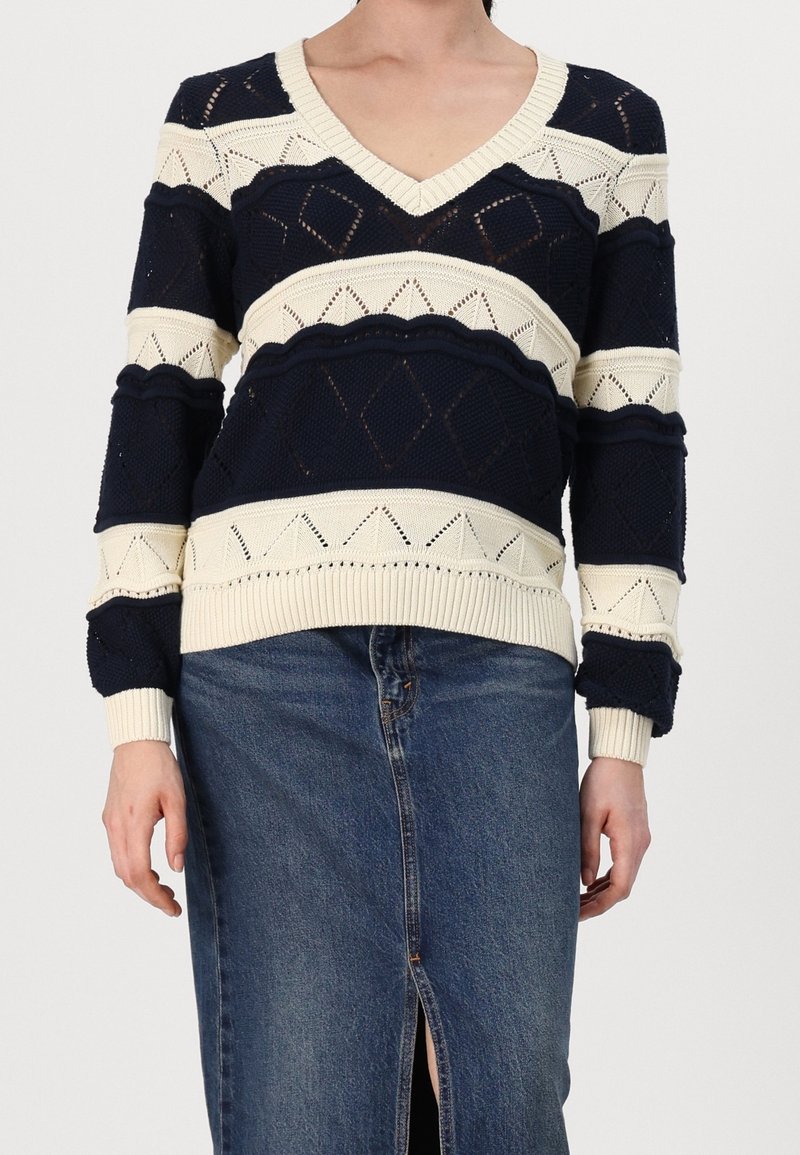 Personne portant un pull en maille à col en V à rayures bleu marine et crème, associé à une jupe en jean mi-longue avec une fente à l'avant.