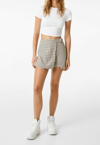 Weißes, gekürztes T-Shirt, kombiniert mit einem beige-braunen Karorock mit seitlichem Knopf und Falten, abgerundet mit weißen High-Top-Sneakers.