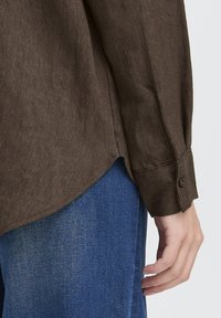 Person in einem braunen Langarmhemd mit geknöpften Manschetten und blauen Denim-Jeans, die Hand entspannt zur Seite.