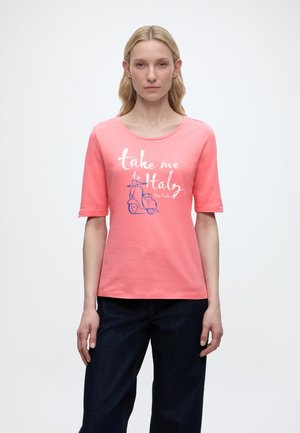Blonde vrouw die een roze T-shirt met korte mouwen draagt met de tekst "breng me naar Italië" en een scooterafbeelding, gecombineerd met donkerblauwe broek, tegen een witte achtergrond.