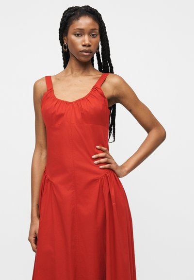 Γυναίκα με μακριά πλεξούδα που φοράει αμάνικο κόκκινο φόρεμα με συγκεντρωμένες λεπτομέρειες, όρθια με το ένα χέρι στη μέση της μπροστά από ένα απλό φόντο.