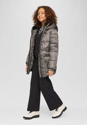 Veste rembourrée grise avec capuche, dotée d'une fermeture éclair et d'une fermeture à pression. Portée par-dessus un pull noir et un pantalon noir large, associée à des bottes blanc cassé.
