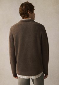 Brauner strukturierter Pullover mit lockerer Passform, langen Ärmeln und leicht abgerundetem Saum, über einem weiß gestreiften Shirt getragen.