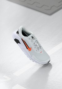 Nike sportsko i lys grå mesh med orange detaljer. Har rund tå, snørebåndsdesign, polstret sål og sorte hæl- og ydersålsdetaljer.