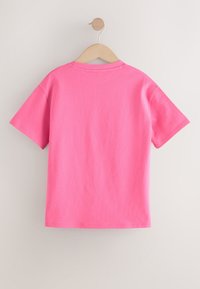 Tričko s krátkym rukávom v jasnej ružovej bavlne, s klasickým okrúhlym výstrihom a hladkou, rovnomernou textúrou, visí na drevenom ramienku.