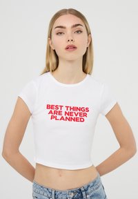 T-shirt bianco a maniche corte e taglio cropped con testo rosso audace che dice "LE MIGLIORI COSE NON SONO MAI PROGRAMMATE". Tessuto di cotone liscio.