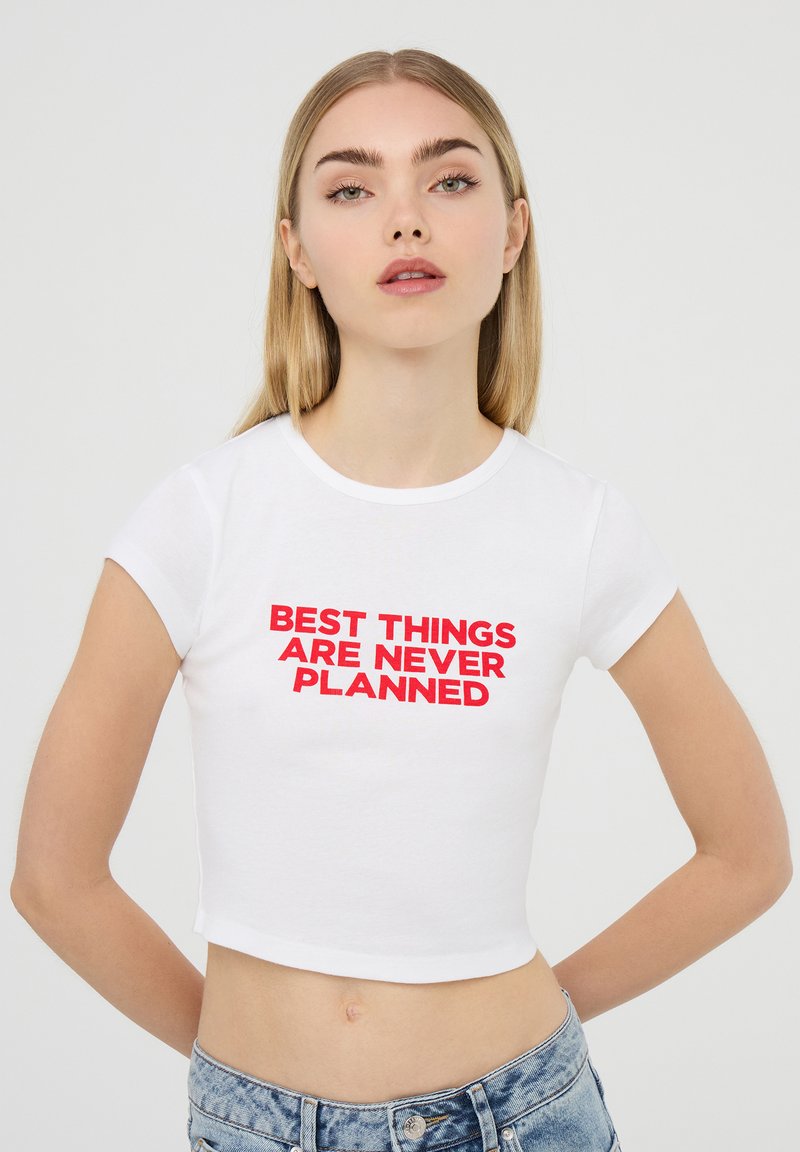 T-shirt bianco a maniche corte e taglio cropped con testo rosso audace che dice "LE MIGLIORI COSE NON SONO MAI PROGRAMMATE". Tessuto di cotone liscio.