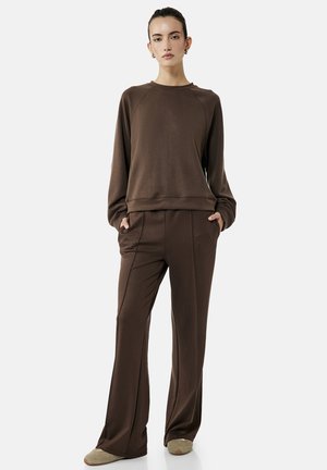 Kvinde iført en mørkebrun langærmet sweatshirt og matchende bukser med vide ben, med hænderne i lommerne, stående på en lys baggrund.