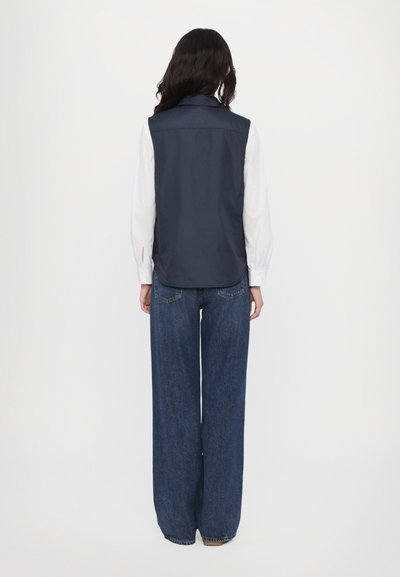 Marineblauw mouwloos vest met een gladde textuur en puntkleur, gedragen over een wit shirt, in combinatie met breedvallende spijkerbroek.