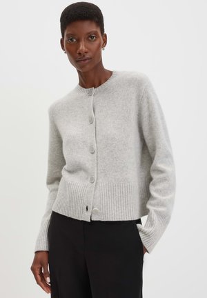 Woman wearing a light gray buttoned cardigan with ribbed hem and cuffs, paired with black pants, standing against a white background.