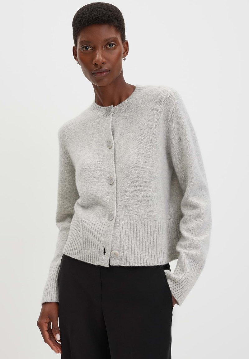 Donna che indossa un cardigan grigio chiaro con bottoni, con orli e polsini a coste, abbinato a pantaloni neri, in piedi contro uno sfondo bianco.
