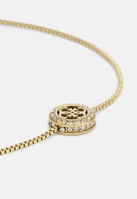 Bracciale d'oro con un design elegante e flessibile. Presenta un ciondolo centrale circolare bordato di strass trasparenti e un motivo intricato.