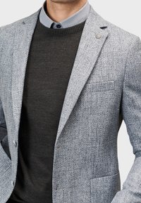 Jasnoszary marynarka o teksturowanej powierzchni z klapami w kształcie notch, z pojedynczym zapięciem na guzik i detalem kieszeni, noszona na ciemnym swetrze i koszuli z kołnierzykiem.