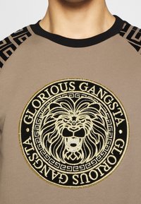 Felpa beige con dettagli neri, caratterizzata da un grande logo di leone ricamato in oro e la scritta "GLORIOUS GANGSTA" in un design circolare.