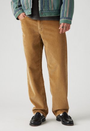 Mężczyzna ubrany w beżowe spodnie sztruksowe, czarne skórzane mokasyny, zieloną kurtkę w paski, z widocznymi tatuażami na prawej ręce.