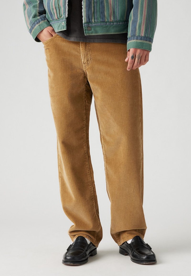 Homme portant un pantalon en velours côtelé beige, des mocassins en cuir noir, une veste rayée verte, avec des tatouages visibles sur la main droite.