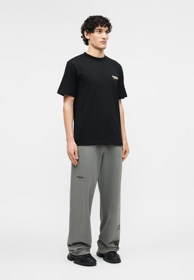 Νέος άνδρας με σγουρά μαλλιά στέκεται φορώντας μαύρο κοντομάνικο T-shirt, γκρι φαρδιά φόρμα με λογότυπα και μαύρα αθλητικά παπούτσια, μπροστά σε απλό φόντο.