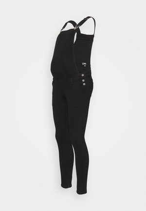 Musta äitiyshaalari, jossa säädettävät olkahihnat, kolme sivupainiketta, etutasku rinnassa ja istuvat kapeat lahkeet valkoista taustaa vasten.