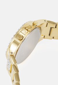 Montre-bracelet en or avec un bracelet texturé, dotée d'un boîtier rond, de trois boutons et d'une lunette ornée de cristaux transparents.