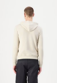 Felpa color beige chiaro con una texture morbida, cappuccio con coulisse e polsini a coste. Indossata con pantaloni corti scuri, mostrando una vestibilità casual e rilassata.