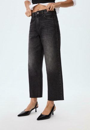 Kvinde iført højtaljede, sorte vaskede straight-leg jeans og sorte spidse slingbackhæle, som retter på sin hvide skjorte.