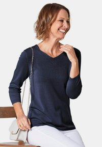 Maglione navy a collo a V con dettagli in pizzo. Presenta maniche lunghe e una vestibilità rilassata. Abbinato a pantaloni bianchi e una borsa leggera.